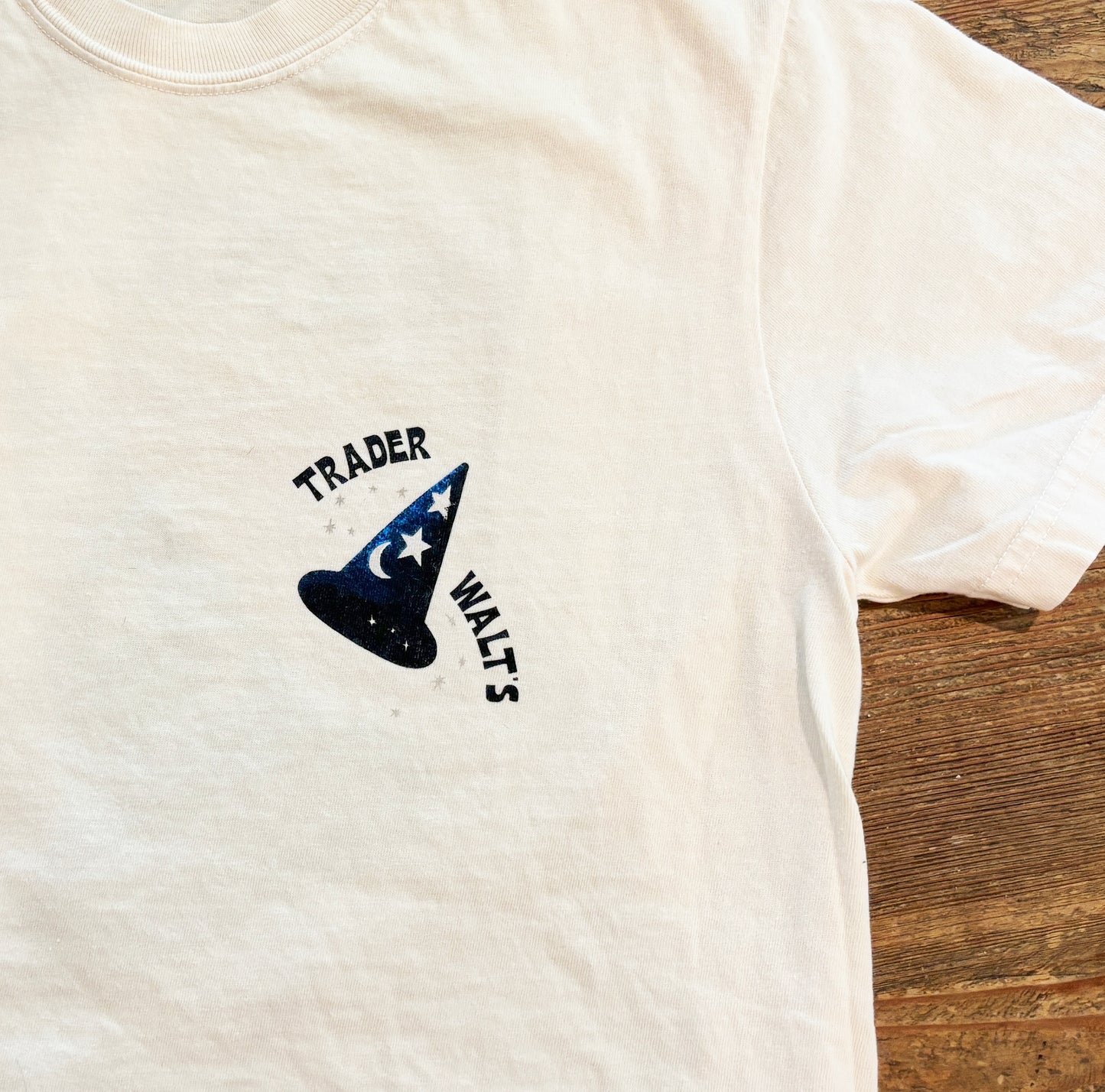 White t-shirt with a blue wizard Mickey-inspired hat graphic and 'Trader Waltz' text on a wooden surface