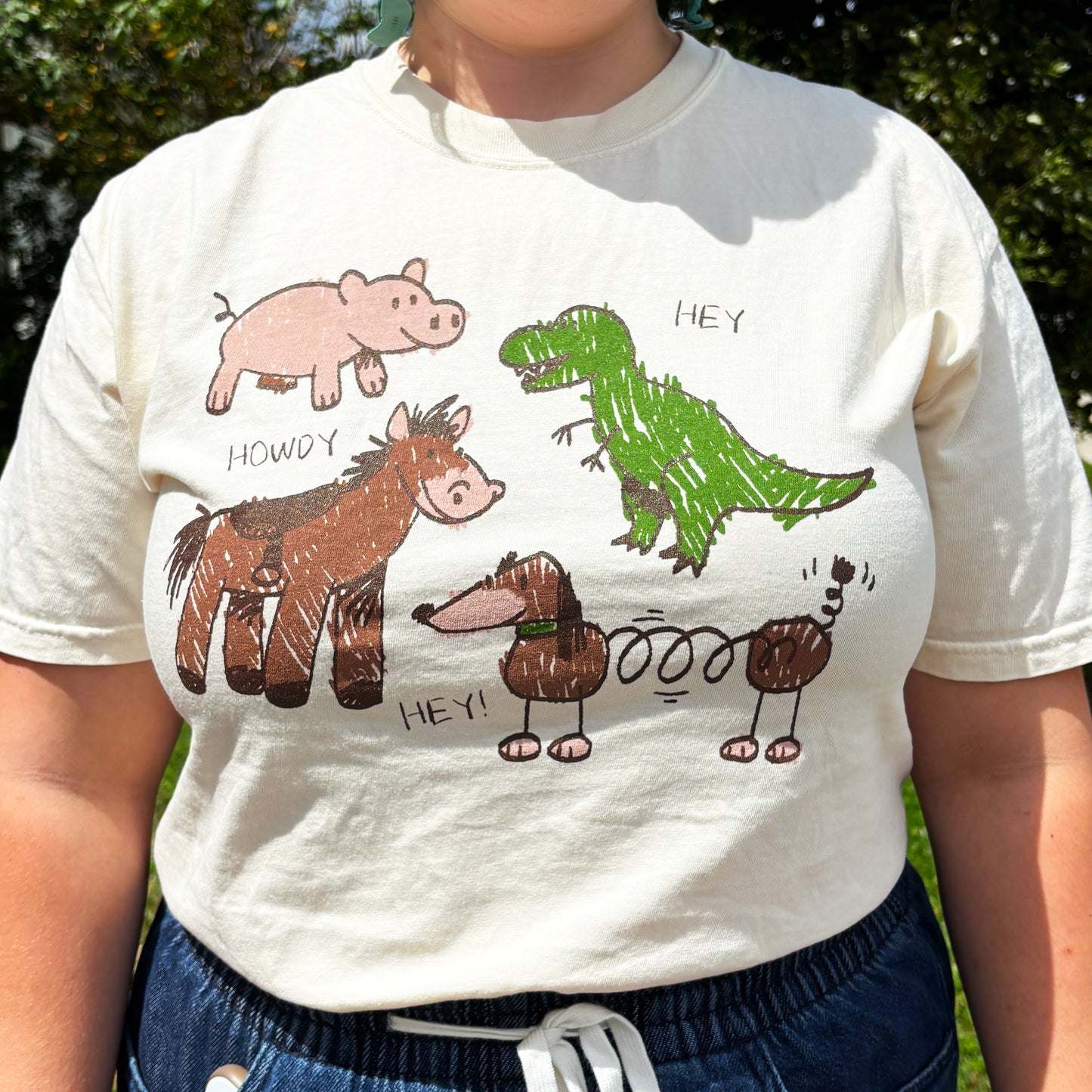 Natural cotton T-shirt featuring crayon-style drawings of a Toy Story Inspired Characters Hamm the pig, woody's horse, Rex the green dinosaur, and Slinky Dog, the springy dog in playful toybox style all saying Hey