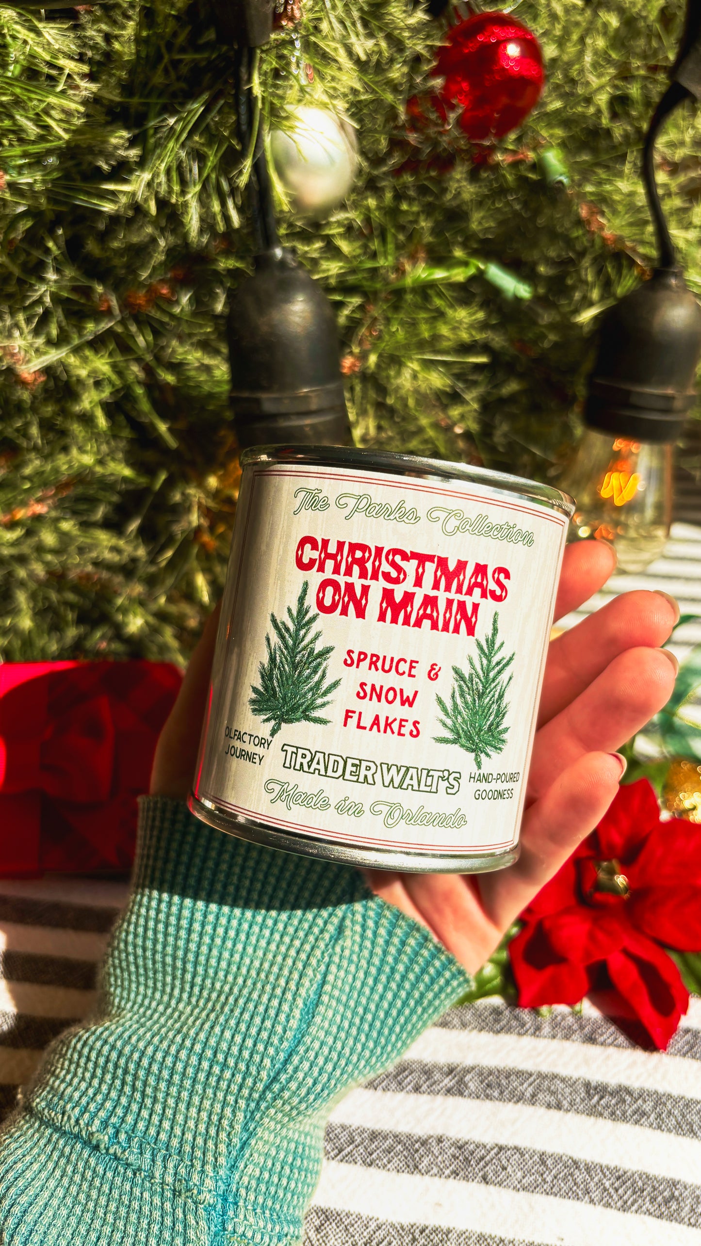 Trader Walt’s ‘Christmas on Main’ candle held in front of a decorated Christmas tree with ornaments and vintage-style holiday lights. Label features spruce branches and text describing a spruce and snowflakes scent.