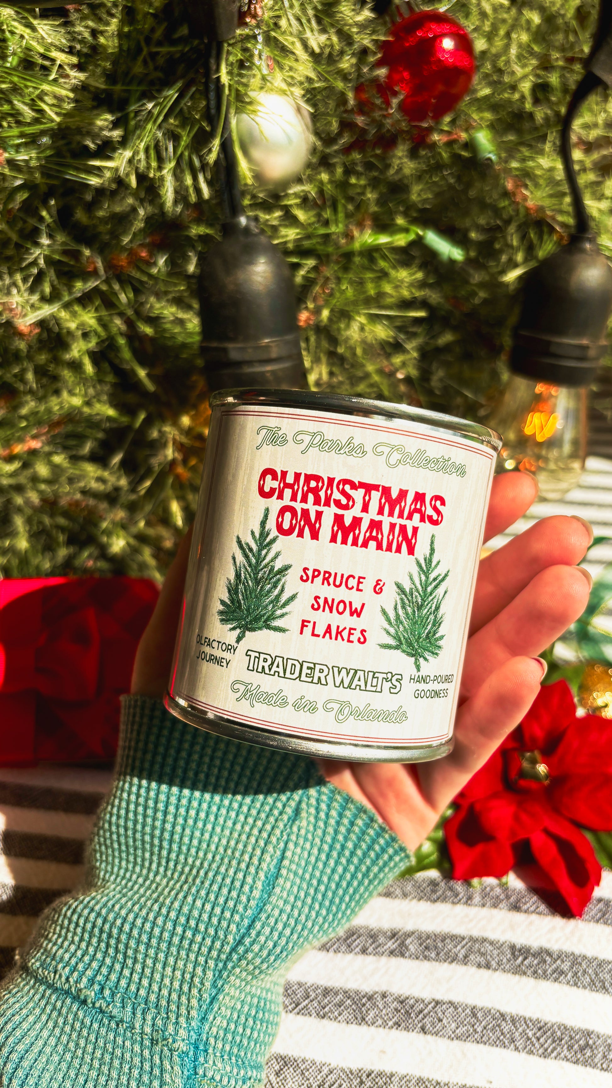 Trader Walt’s ‘Christmas on Main’ candle held in front of a decorated Christmas tree with ornaments and vintage-style holiday lights. Label features spruce branches and text describing a spruce and snowflakes scent.