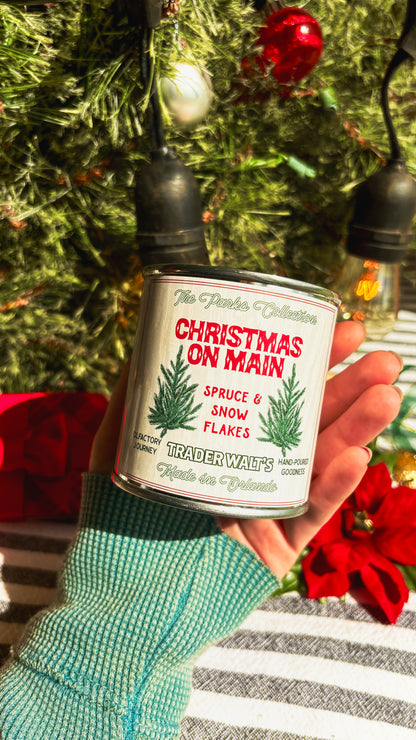 Trader Walt’s ‘Christmas on Main’ candle held in front of a decorated Christmas tree with ornaments and vintage-style holiday lights. Label features spruce branches and text describing a spruce and snowflakes scent.