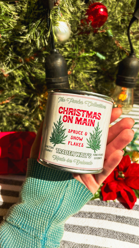 Trader Walt’s ‘Christmas on Main’ candle held in front of a decorated Christmas tree with ornaments and vintage-style holiday lights. Label features spruce branches and text describing a spruce and snowflakes scent.