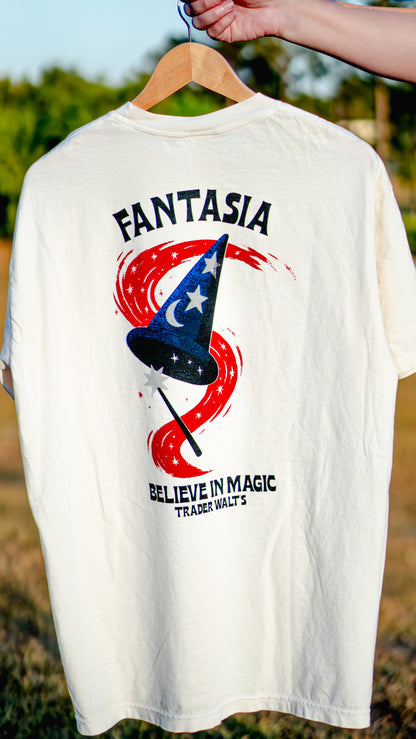 The image shows a cream-colored T-shirt with the word 'FANTASIA' written across the back in large red letters, featuring a graphic of a wand with a blue star and red accents. The T-shirt is displayed on a hanger, and part of a person's hand is visible at the top of the frame.