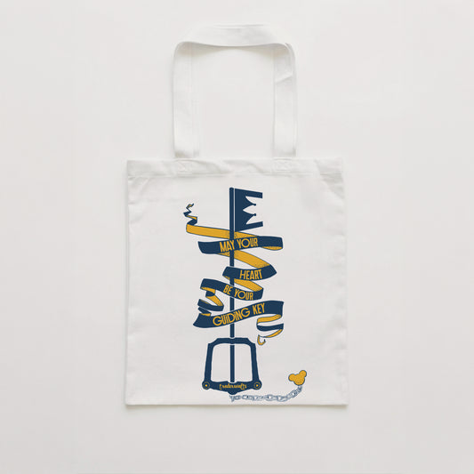 White tote bag with a screen printed navy and gold design of a keyblade wrapped in a flowing ribbon banner that reads May Your Heart Be Your Guiding Key
