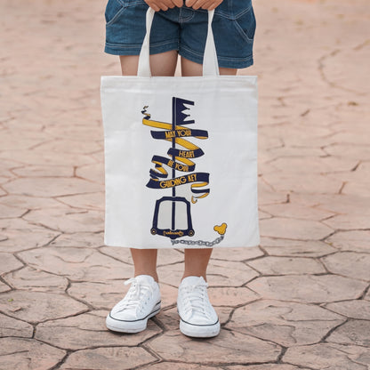 Girl standing with a White tote bag with a screen printed navy and gold design of a keyblade wrapped in a flowing ribbon banner that reads May Your Heart Be Your Guiding Key